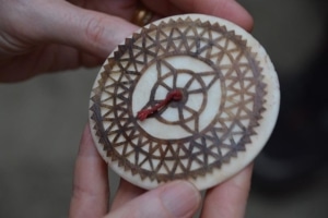 A small, round, carved ornament with brown detail showing a lacelike pattern which resembles a compass