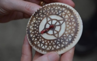 A small, round, carved ornament with brown detail showing a lacelike pattern which resembles a compass