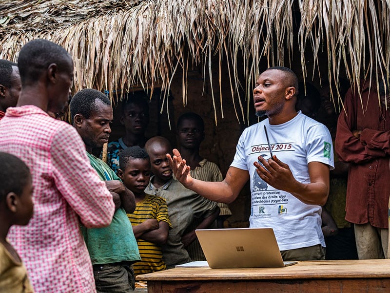 person talking to a group of people with a computer Infront of him and a object in his hand