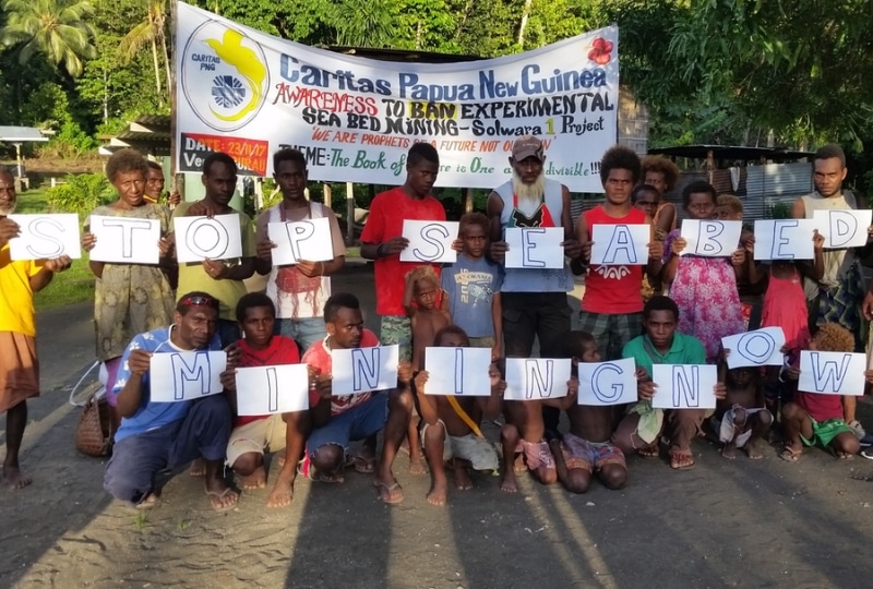 A group of men, women, and children in Papua New Guinea hold up individual signs that together read ‘STOP SEABED MINING NOW.’ Behind them is a banner from Caritas Papua New Guinea raising awareness to ban experimental seabed mining under the Solwara 1 Project.