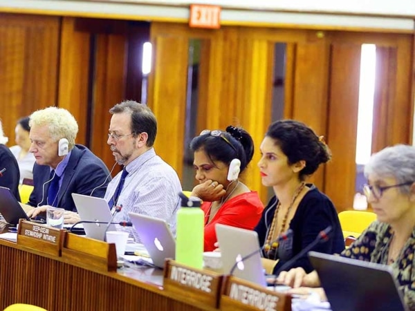 Five people sitting at a long wooden table with their laptops, some of them wearing white devices over their ears, there are wooden signs in front of the people with the names of their organisations and one of them appears to be watching something happening on the other side of the room