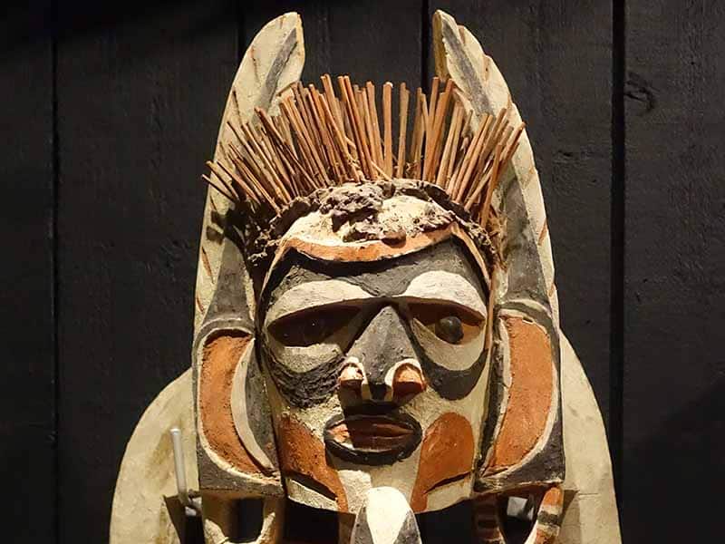 A close-up of the face of a painted wooden Malagan carving with strong use of shapes (circular eyes, triangular nose), two long feather-like structures either side of the head and a bunch of sticks growing out of the head