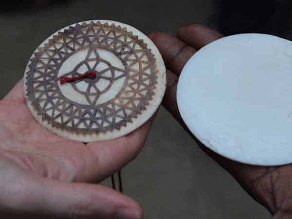 Two hands, one light-skinned and one dark-skinned, holding two round objects. In the left hand the object is wooden and appears to have a piece of red thread tied in a knot through the middle, and a dark symmetrical pattern around it. The other round object is plain white with no markings.