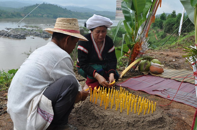 On a ridge next to a wide river, a man and a woman light an arrangement of around thirty yellow candles which have been pushed into an oval-shaped mound of sand.
