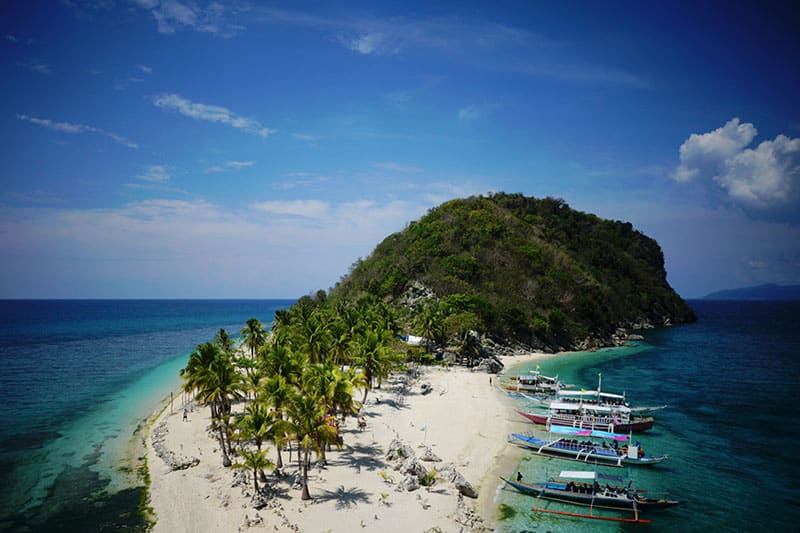 An island with a white sand beach and palm trees lined with colourful boats, leading to a small hill covered in green shrubbery.