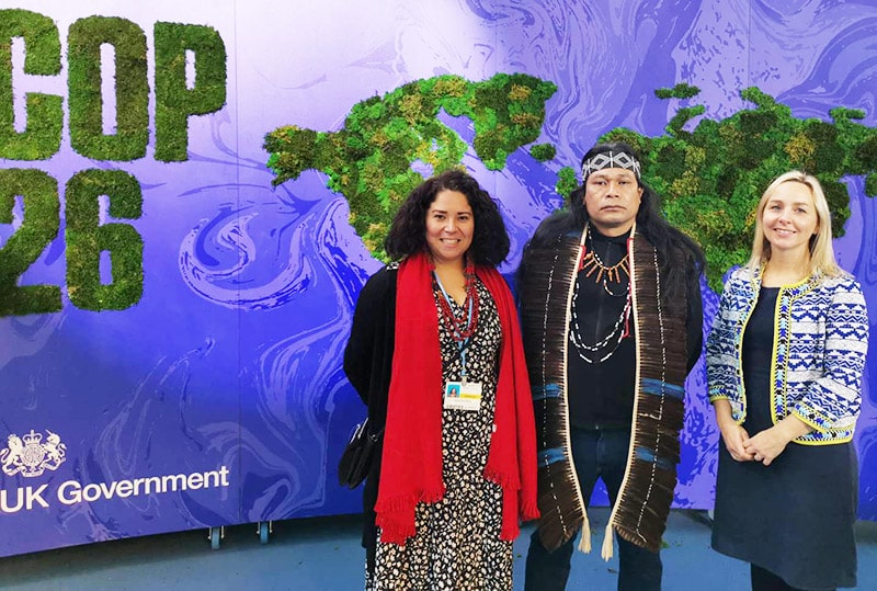 Three people standing and smiling in front of a purple banner with a world map the words 'COP 26' and 'UK Government'