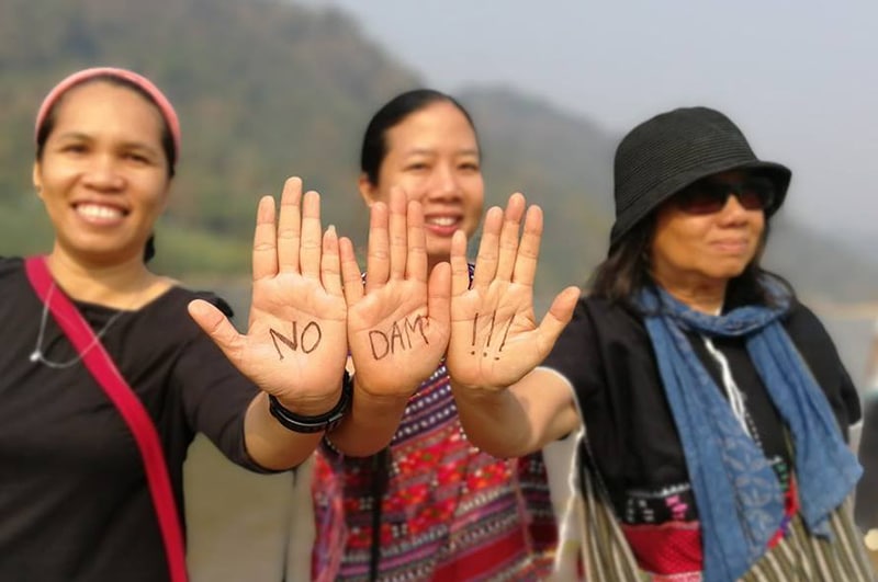 Three women stand in front of a river, each holding up one hand with each hand touching the edges of the next. One each palm is text, and altogether it reads NO _ DAMS _ !!!
