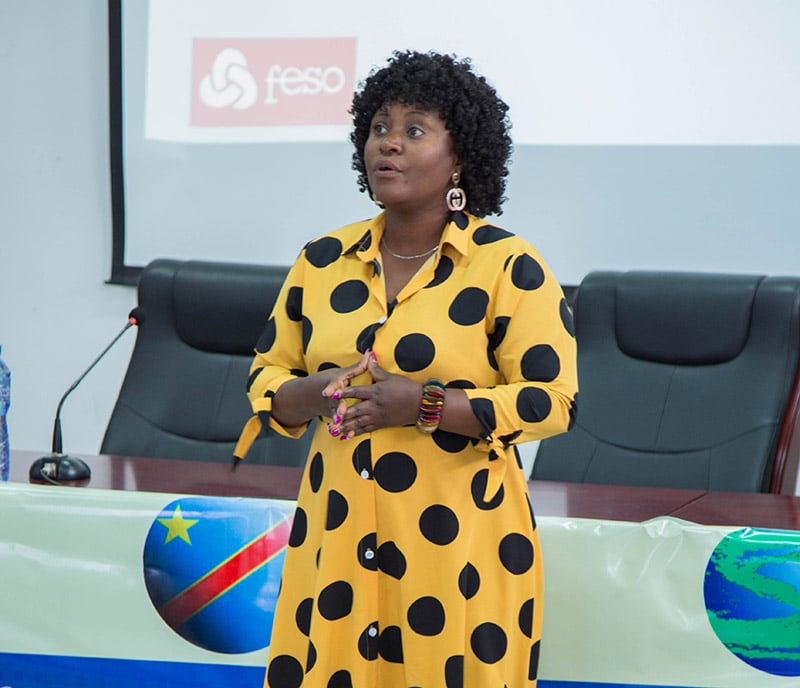 A lady in a yellow dress with black spots is speaking at a podium with a presentation behind her with the FESO logo.