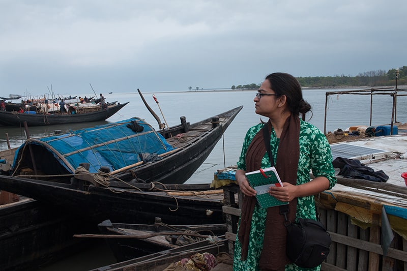 Alifa standing at a harbour with lots of fishing boats, notebook in hand.