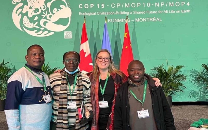 Synchronicity Earth Co-founder Jessica Sweidan with Jean-Christophe Bokika (MMT), Dr Bihini Won wa Musiti (SE Congo Basin Affiliate), and Joseph Itongwa (ICCA Consortium) standing together in front of flags and a COP15 banner at the Convention on Biological Diversity COP15 in Montreal 