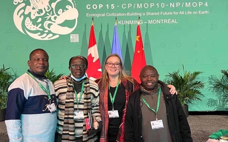 Synchronicity Earth Co-founder Jessica Sweidan with Jean-Christophe Bokika (MMT), Dr Bihini Won wa Musiti (SE Congo Basin Affiliate), and Joseph Itongwa (ICCA Consortium) standing together in front of flags and a COP15 banner at the Convention on Biological Diversity COP15 in Montreal 