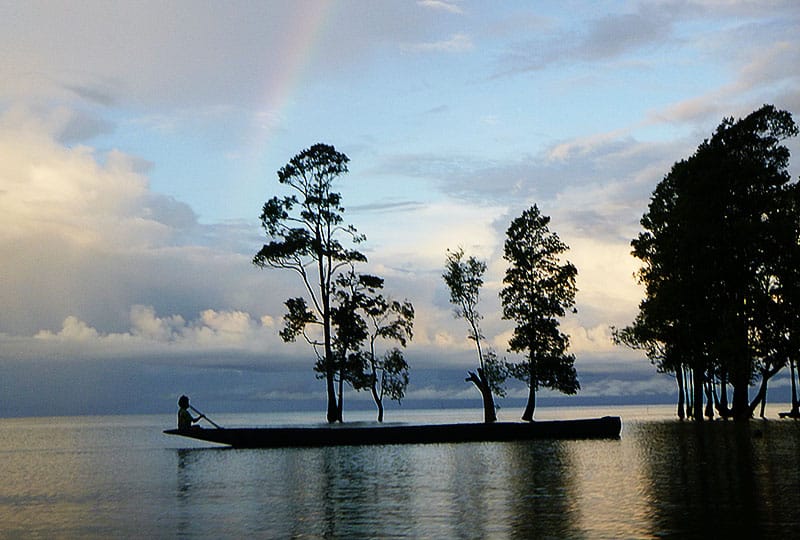 A narrow fishing boat silhouetted on a coastline with trees, a calm sea and rainbow in a cloudy sky.