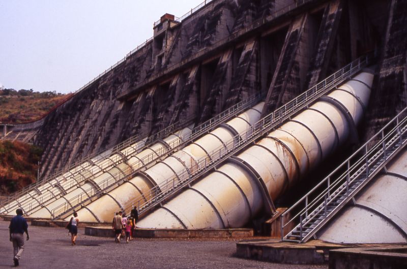 View of the wall of Inga 1 hydropower dam, with people walking towards it. The wall is many times taller than a person, with massive tubes projecting upwards.