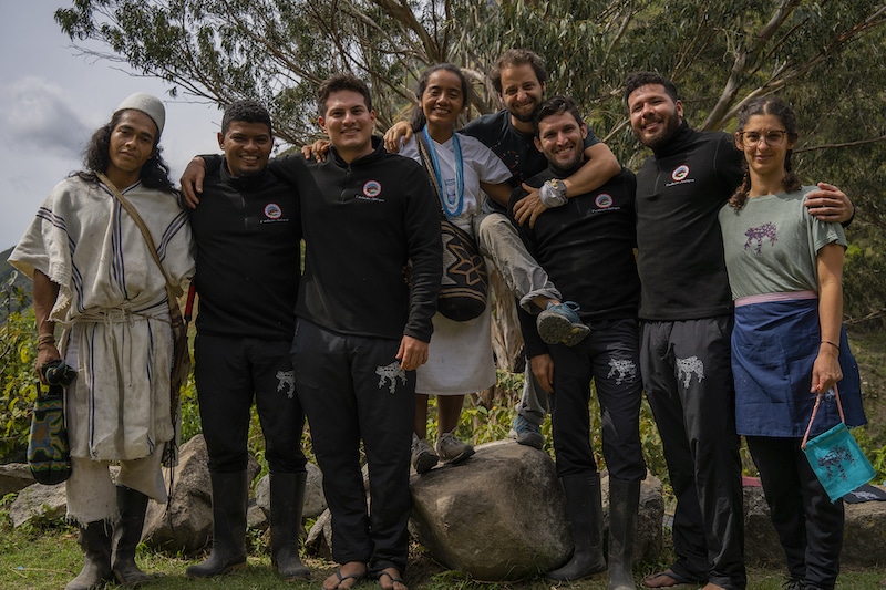 Eight people standing in a row and smiling. Some wear black t-shirts with the Fundacion Ateloupos logo, others wear traditional garments. 