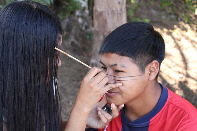 Someone is painting someone else's face with thin black lines horizontally across their nose and cheekbones.