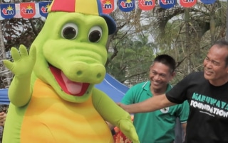A giant crocodile mascot wearing a sideways baseball cap waving, with two delighted bystanders
