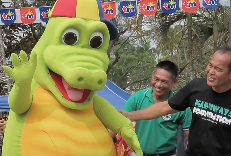 A giant crocodile mascot wearing a sideways baseball cap waving, with two delighted bystanders