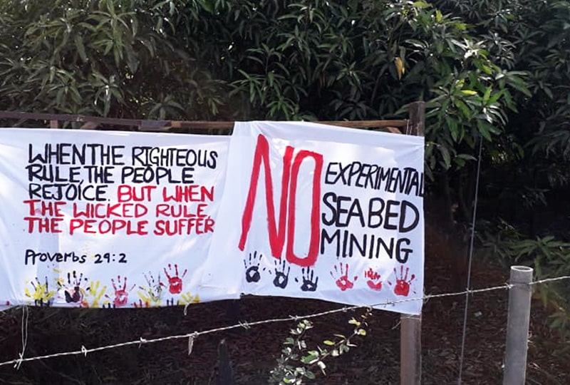 A banner is placed in front of a background of a trees. The banner reads: 'When the righteous rule, the people rejoice. But when the wicked rule, the people suffer. Proverbs 29:2. NO experimental seabed mining'. 
