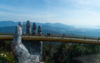 A large sculpture of a human hand is holding a footbridge up with people crossing it and forests below