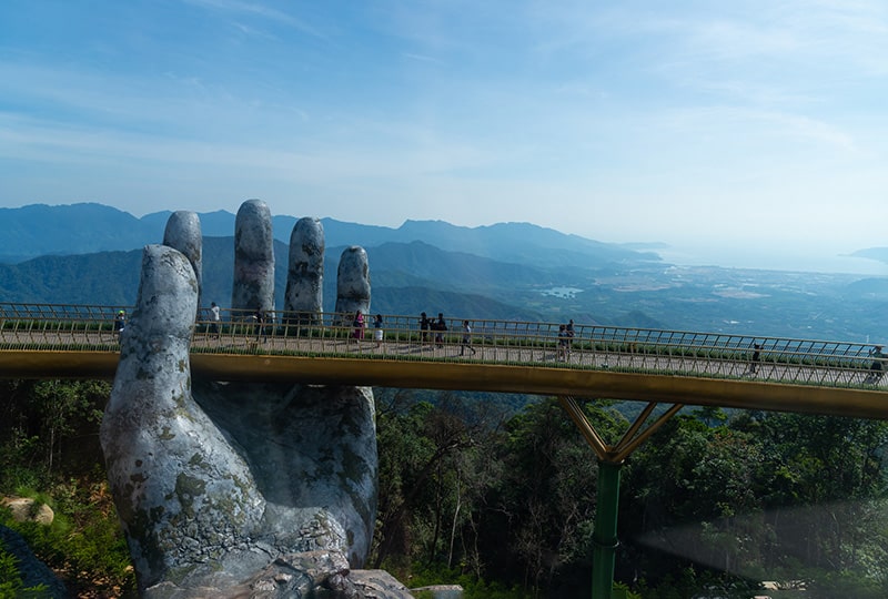 A large sculpture of a human hand is holding a footbridge up with people crossing it and forests below