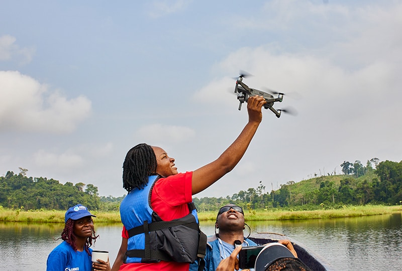 Merline stands in a boat on the river and holds up a drone towards the sky.