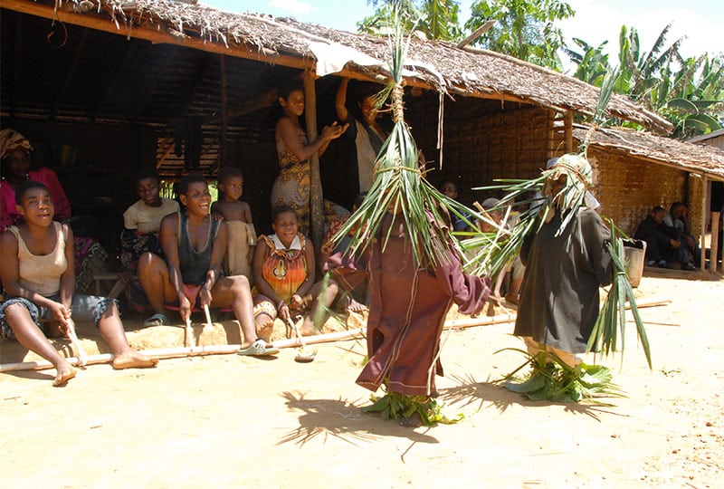 A local community sit outside in the shade, with some in Bagyeli Indigenous dress made of leaves. 