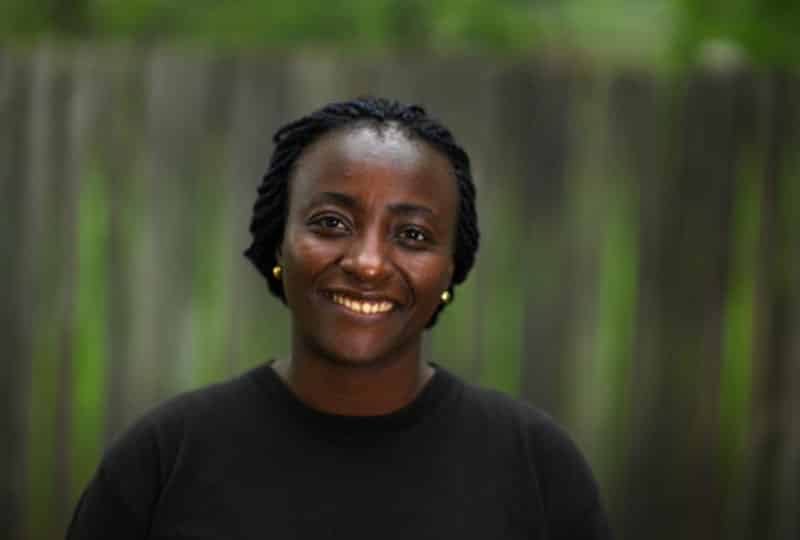 Marina smiles wearing a black jumper against a blurred green background. 