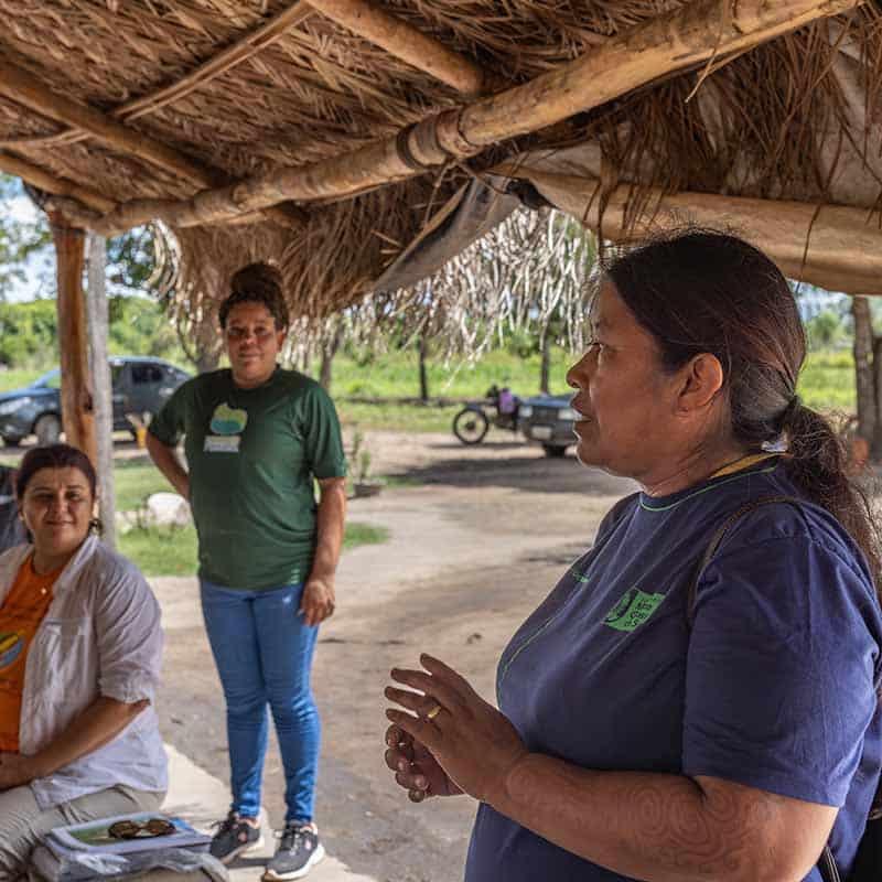 Three women in conversation under a straw roof at an outdoor community meeting in the Pantanal, Brazil, facilitated by MUPAN