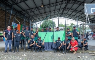 Group photo of uniformed individuals holding spears, standing in front of a green and black flag under a metal-roofed structure. Two individuals not in uniform stand on either side of the group, one in traditional attire.