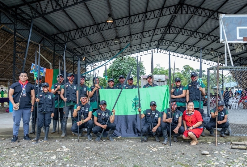 Group photo of uniformed individuals holding spears, standing in front of a green and black flag under a metal-roofed structure. Two individuals not in uniform stand on either side of the group, one in traditional attire.