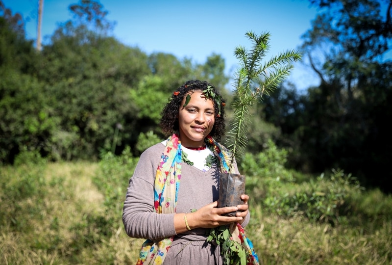 Woman holding a tree sapling in a field, wearing a colourful scarf and a crown of leaves.