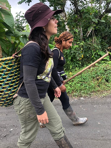 Two young women walk side by side with baskets on their back containing seedling for reforestation