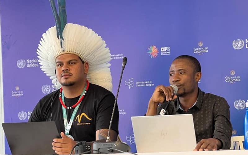 Two Indigenous men sit on panel, one speaking into a microphone