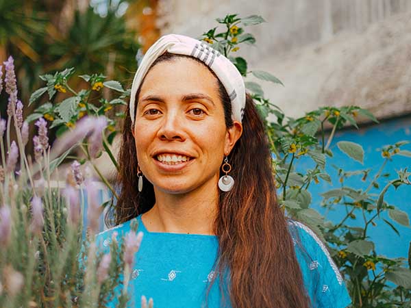 A woman with long dark hair and a blue top stands among flowers