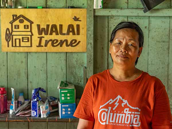 A woman dressed in an orange t-shirt stands inside a wooden building with a sign in the background, looking at the camera