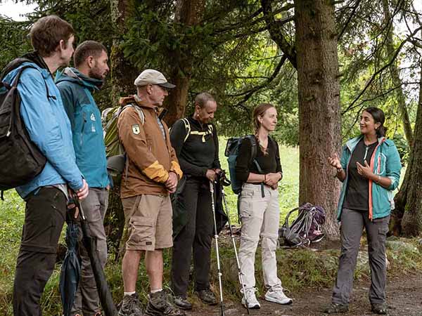 A group of people stand in an Alpine forest listening to a woman speaking