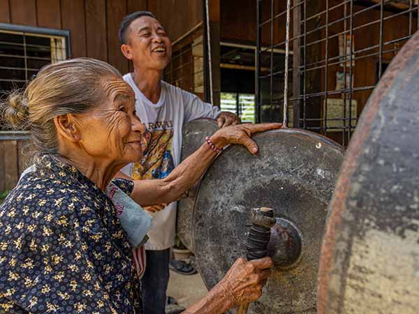 An elderly female villager plays a gong with a man smiling in the background