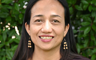 Image of Kate Mana Galido smiling at a camera. She is pictured from the shoulders up in front of a green bush.