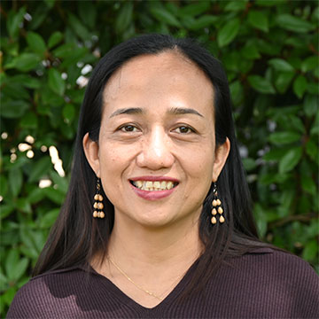 Image of Kate Mana Galido smiling at a camera. She is pictured from the shoulders up in front of a green bush.