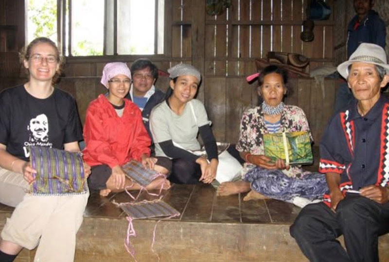 Kate pictured with Datu Amay Mantangkilan, his wife, and others. They are sat on the floor and hold textiles in a wooden home.