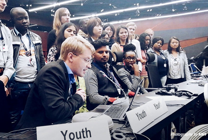 Image of Christian speaking into a microphone at a conference desk. Plaques reading ‘youth’ are placed in front of him. A large group stand behind him.