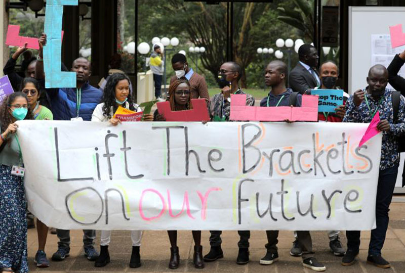 Image of GYBN members holding a large banner reading ‘Lift the brackets on our future’ while other members hold multicoloured brackets made of cardboard.