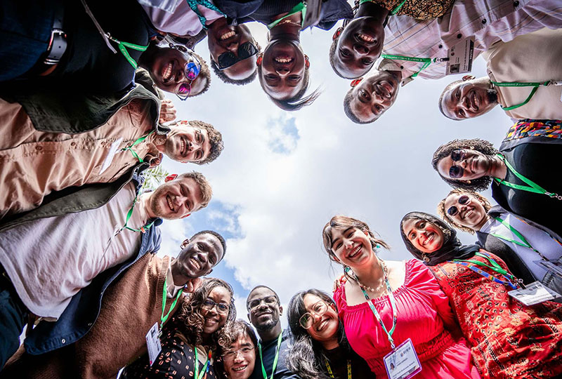 A group of 16 young people standing in a circle, smiling down at a camera that has been placed on the floor.