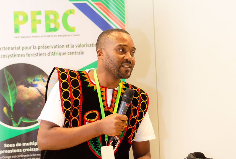 Image of Aristide Chacgom holding a microphone. A poster with information about the CBFP 20th meeting of the parties is in the background, written in French.