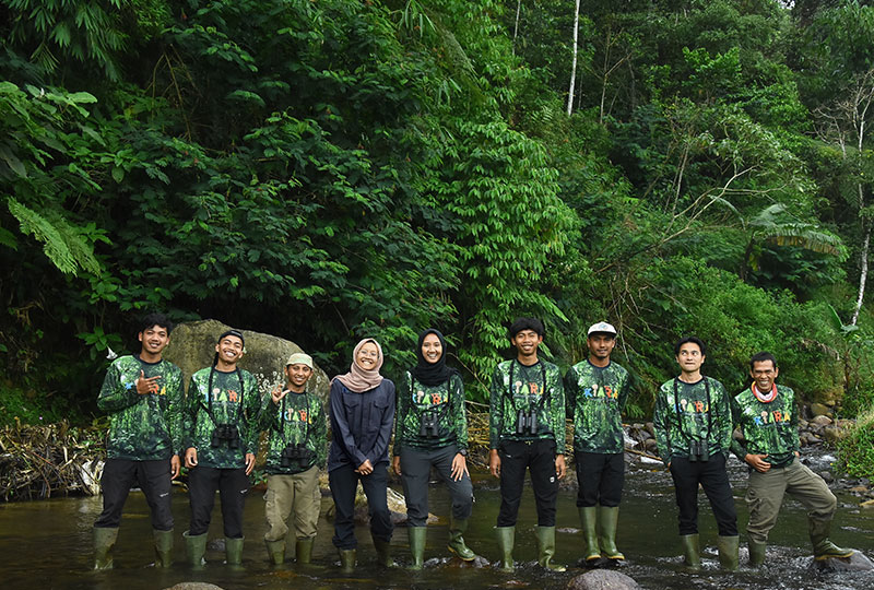 Image of nine people standing in a river in the rainforest – the KIARA team. They all wear green rainboots on their feet and are facing the camera smiling.