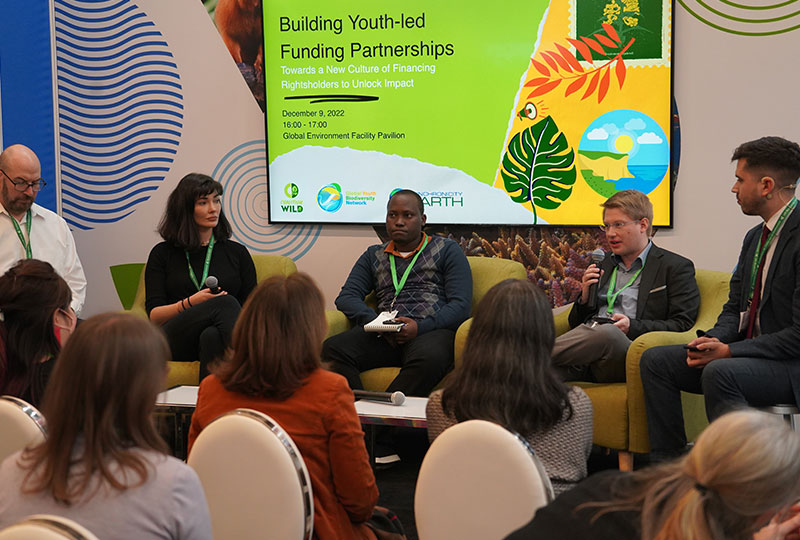 An audience watches a panel with five people. The presentation screen behind reads: 'Building Youth-led Funding Partnerships'