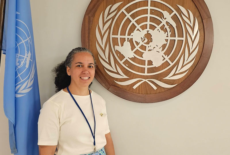 Image of Kathryn Audroing stood in front of a plaque with the logo of the United Nations on it.