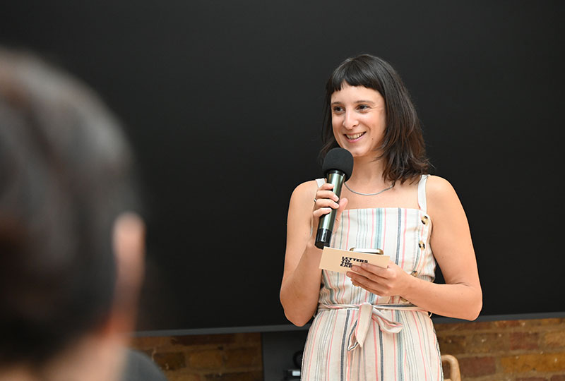 Image of a woman holding a microphone in one hand and a postcard reading Letters to the Earth in the other. She wears a vertically-striped jump suit.