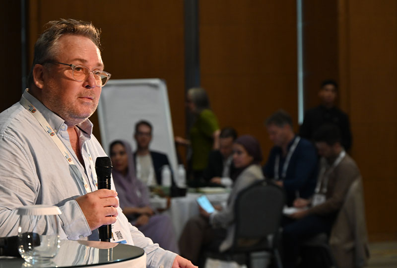 Image of Mike holding a microphone, sitting on stage and addressing a group of people.
