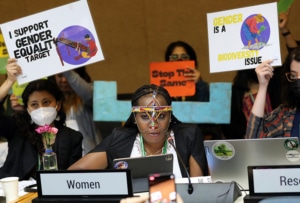 Image of Sharon Ruthia speaking into a microphone at a conference. She sits in front of caucus members holding up signs and wearing medical masks.